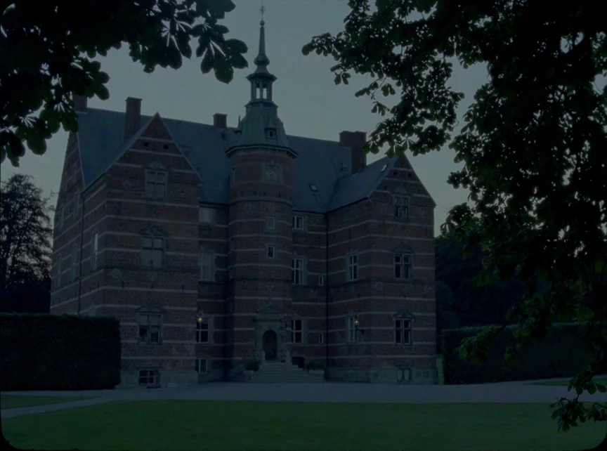 Jauja (2014) movie still: establishing — A historic brick manor house with a central tower stands in a grassy park. Dark trees frame the structure…