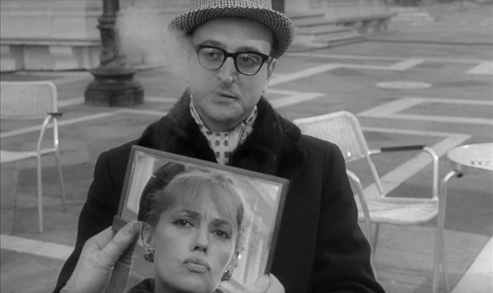 Eva (1962) movie still: medium — A man stands in an outdoor plaza with light paving stones and metal chairs. The man holds up a handheld mirror…