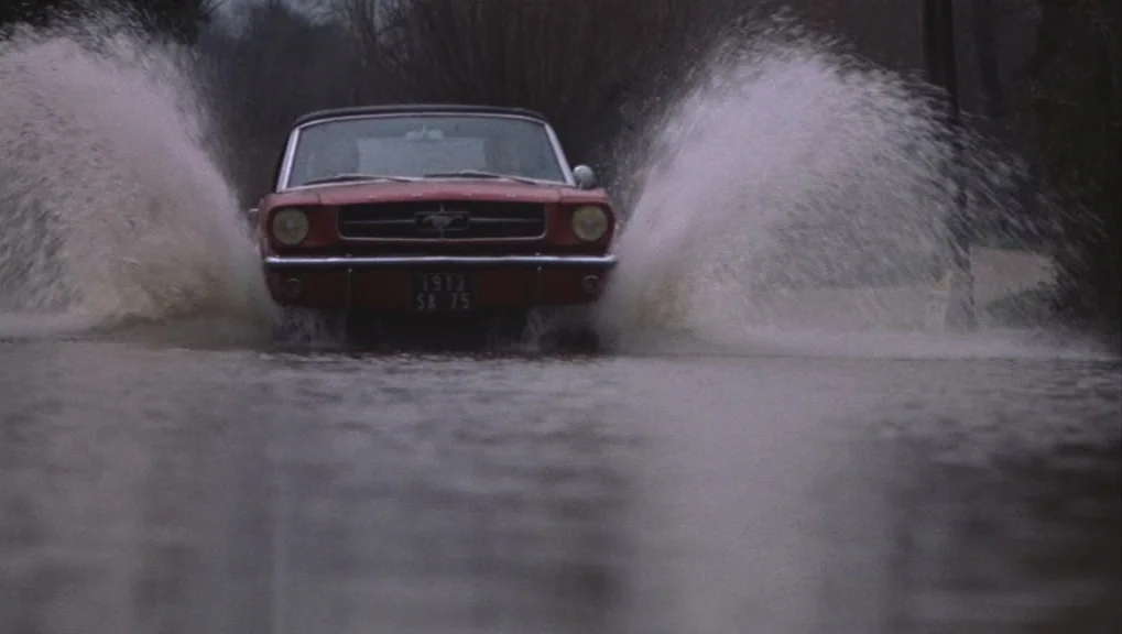Un Homme et Une Femme (1966) movie still: medium — A classic red convertible driving through a flooded, tree-lined asphalt road. Large splashes of…