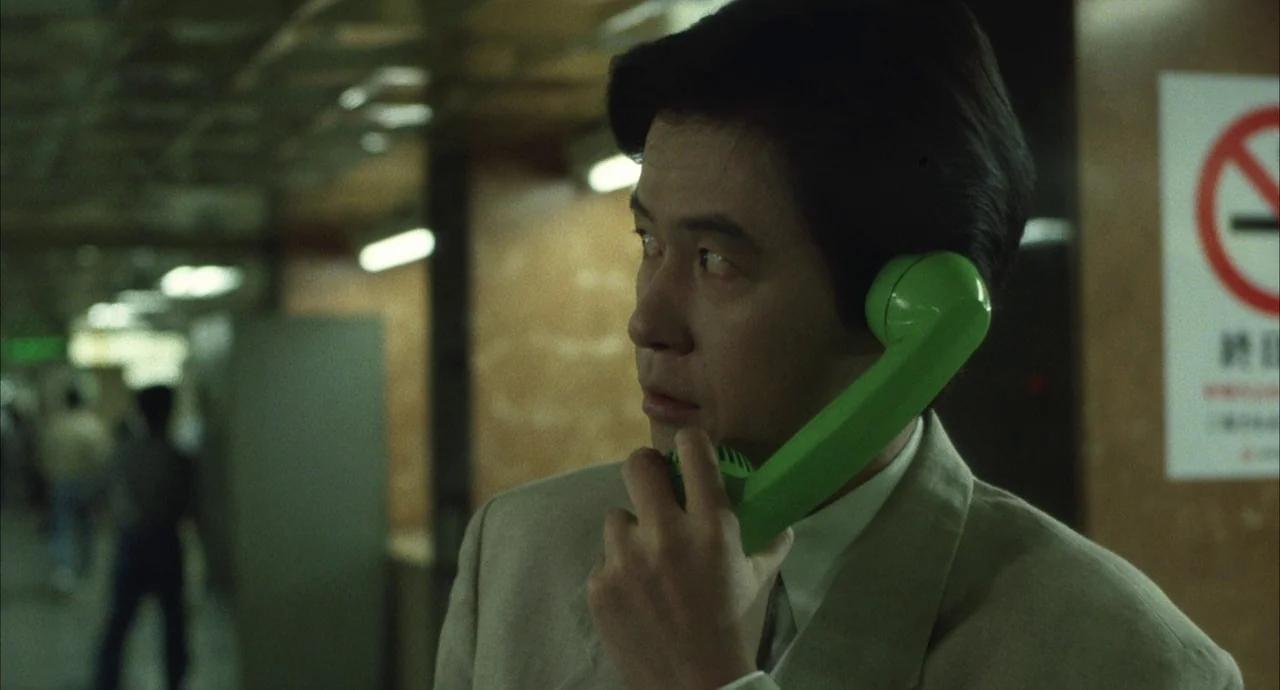 The Discarnates (1988) movie still: medium — A man in a beige suit stands in a tiled subway concourse area. He is holding a bright green public…