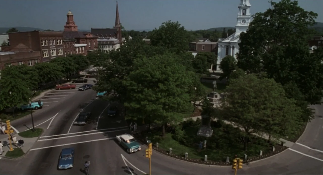 Jumanji (1995) movie still: establishing — A town square features a central park area surrounded by brick buildings and a church. Several vintage…