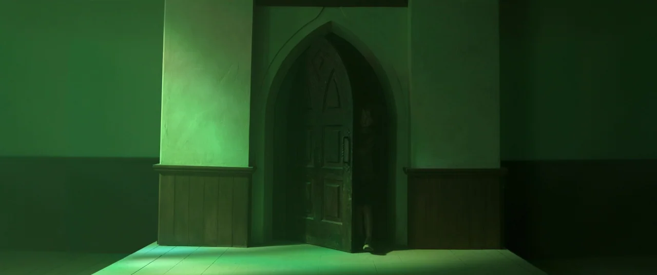 In The Tall Grass (2019) movie still: wide — A dimly lit hallway features a large wooden arched door in the center. The surrounding walls are…