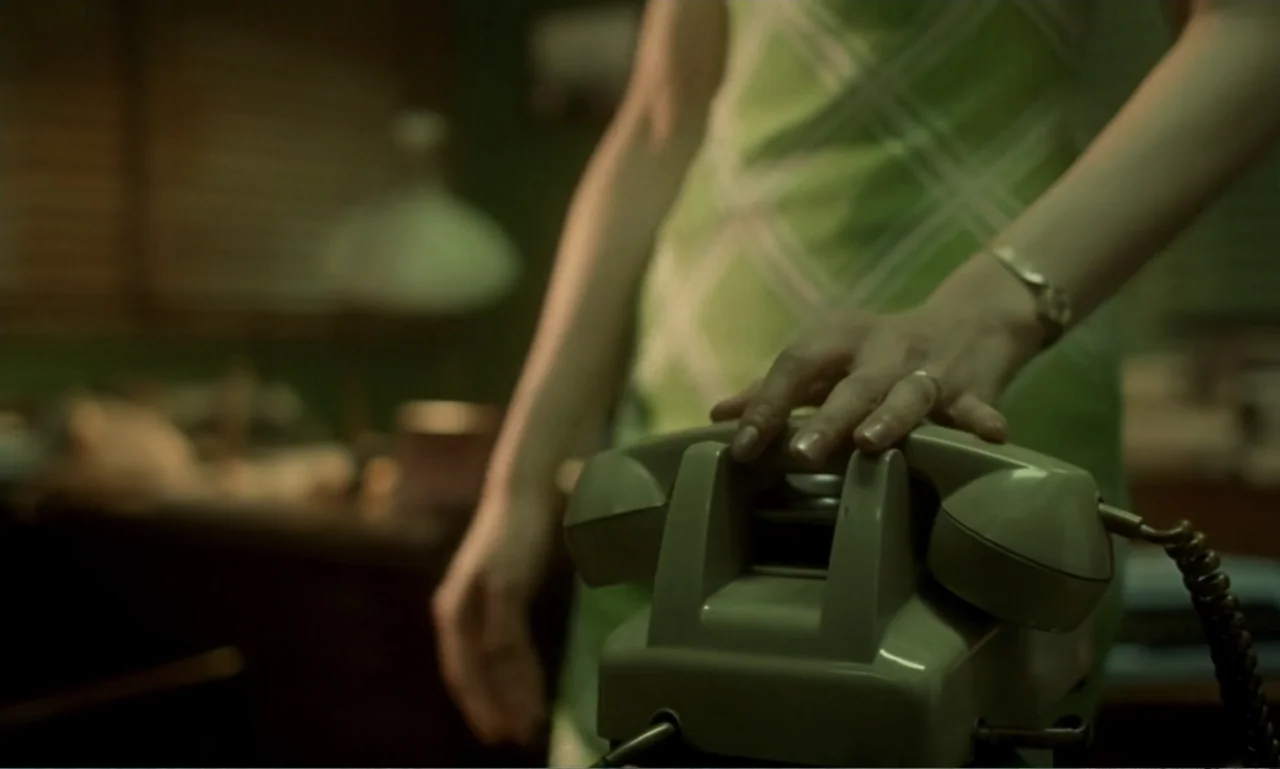 In the Mood for Love (2000) movie still: medium — A dimly lit room contains a desk with a vintage rotary telephone. A person wears a green patterned…