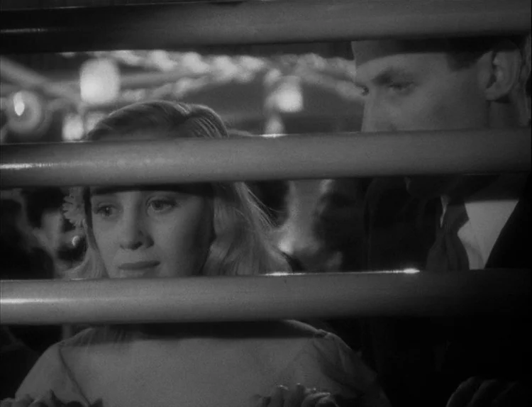 Music in Darkness (1948) movie still: medium — Two people are positioned behind horizontal metal bars inside a dark passenger compartment. The woman…