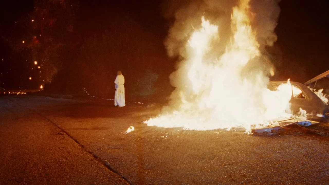 The Sect (1991) movie still: wide — A dark, paved road at night features a car engulfed in flames. A lone figure in a white robe stands nearby…