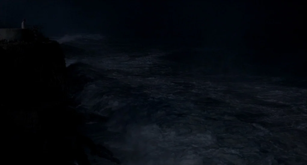 Anatomy of Hell (2004) movie still: wide — A dark, rugged cliff overlooks a churning, turbulent sea at night. A lone figure stands on the elevated…