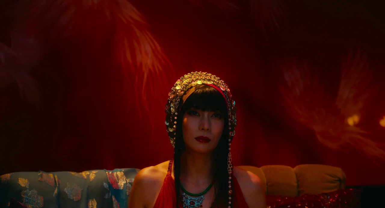 xxxHolic (2022) movie still: medium — A room with deep red walls decorated with faint repeating feather or wing patterns. A woman wearing an ornate…