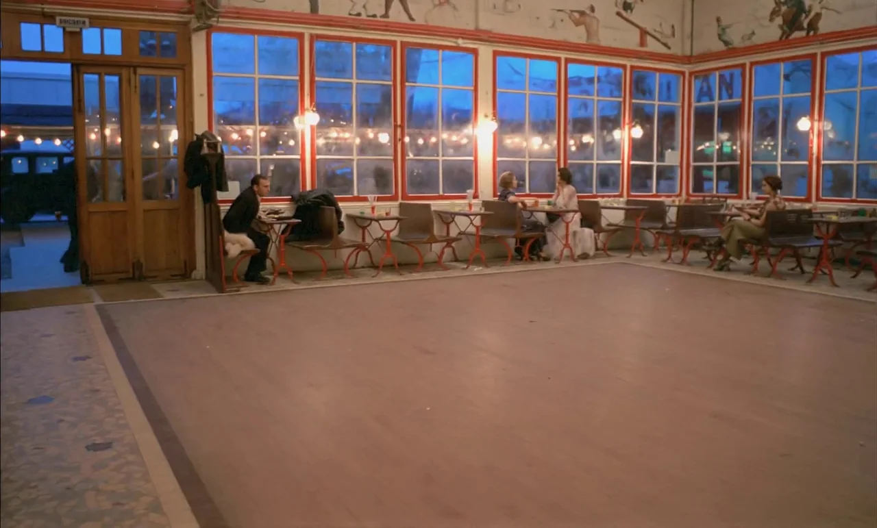 The Conformist (1970) movie still: wide — A spacious dance hall features a large wooden floor and red-framed window panes. Several individuals sit…