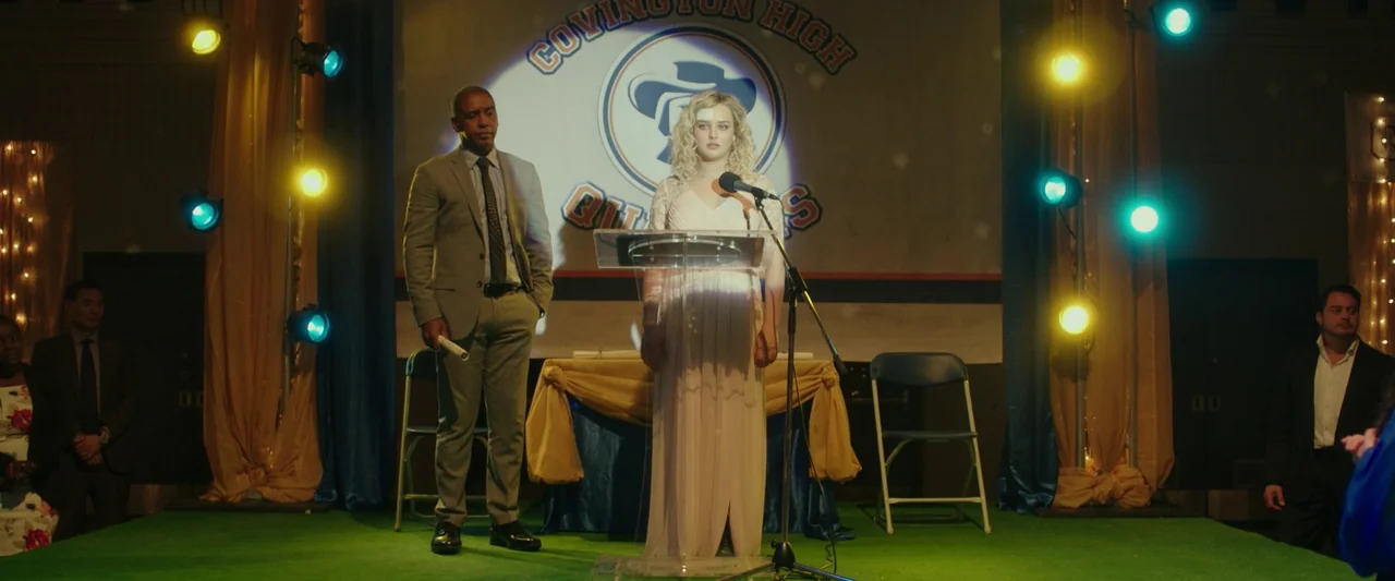 Spontaneous (2020) movie still: fullbody — A decorated high school stage with a large banner depicting the school logo behind. A young woman speaks…