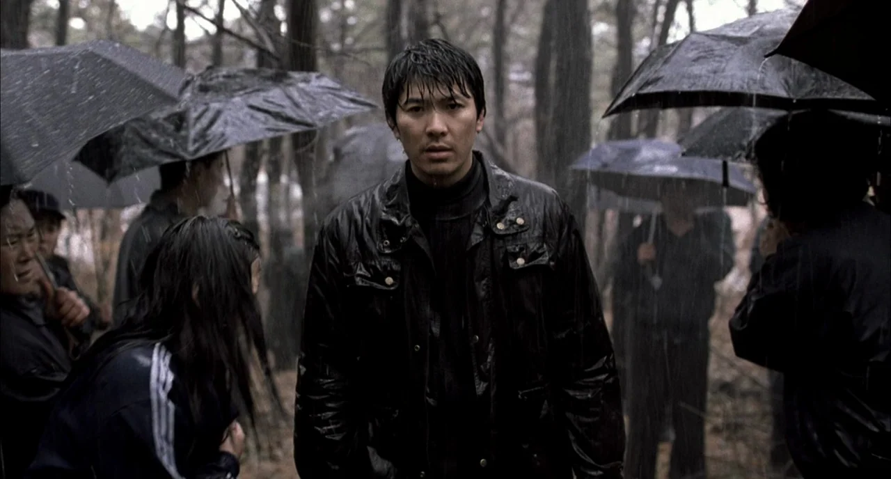 Memories of Murder (2003) movie still: medium — A group of people standing in a wet, gloomy woodland area during a rainstorm. The central man stands…