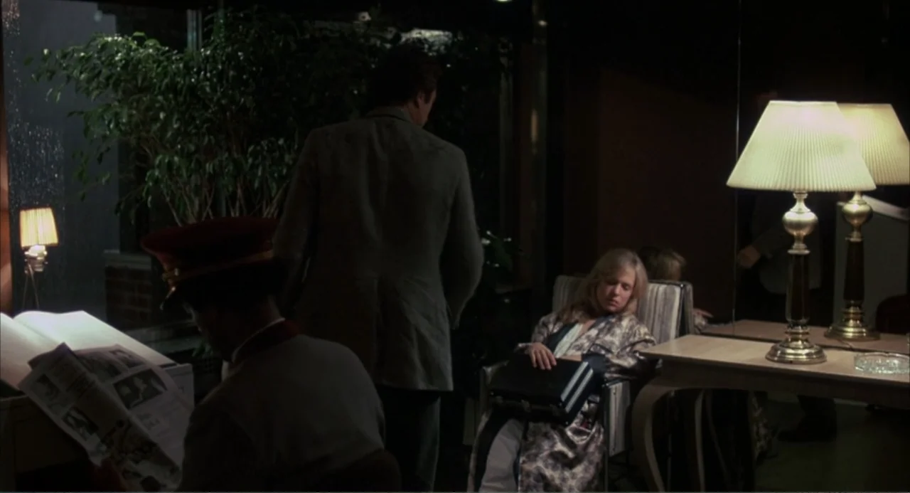 F/X (1986) movie still: medium — A dimly lit hotel lobby features dark wood paneling and large indoor potted plants. A man in a suit stands between…