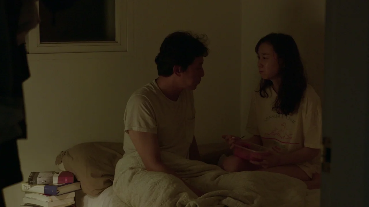 Yourself and Yours (2016) movie still: medium — A dimly lit room contains a bed where two people sit facing each other. The woman holds a plastic…