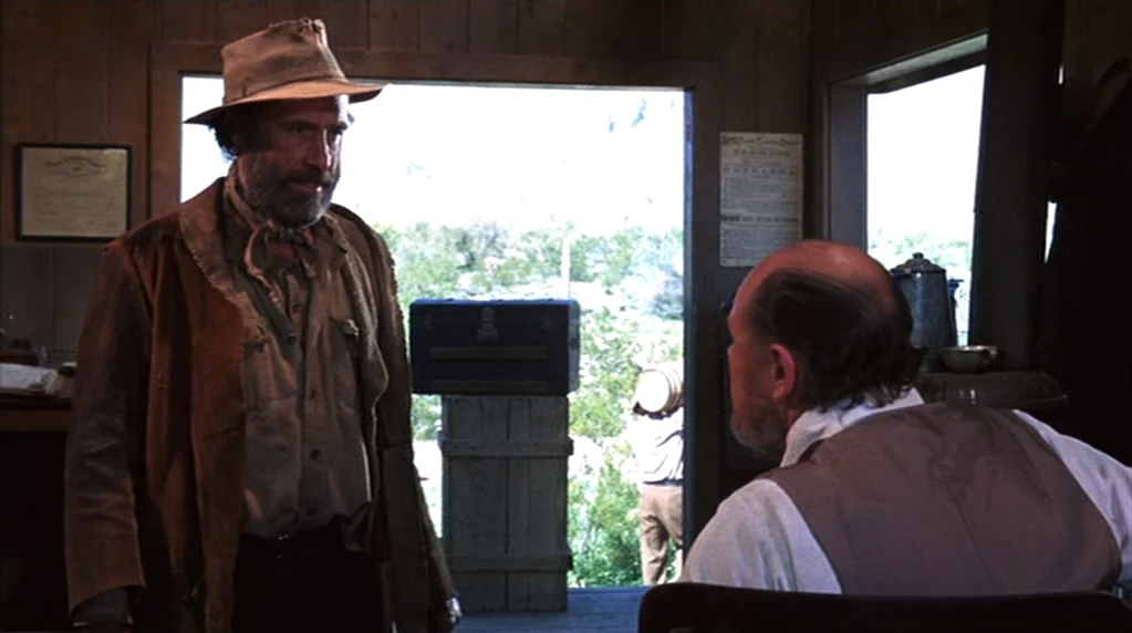 The Ballad of Cable Hogue (1970) movie still: medium — A rustic cabin interior features wood paneled walls and a doorway to a desert exterior. A man…