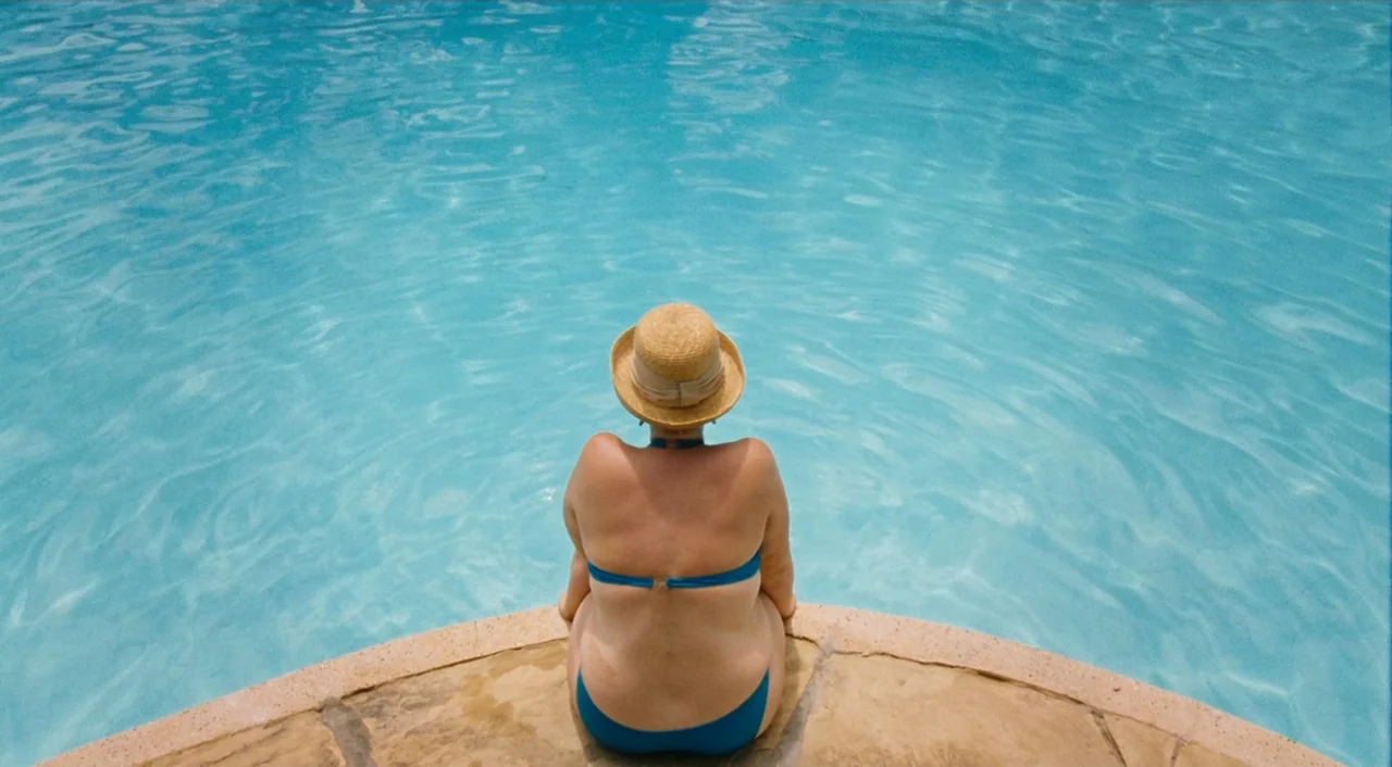 Paradise: Love (2012) movie still: medium — A woman sits at the curved edge of a bright blue swimming pool. She is wearing a straw hat and looks out…
