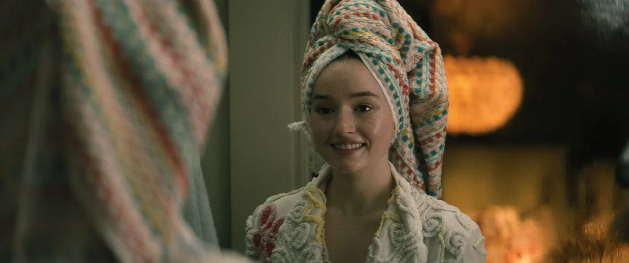 No One Will Save You (2023) movie still: closeup — A woman stands in a bathroom looking at a mirror reflection. She is wearing a multi-colored towel…