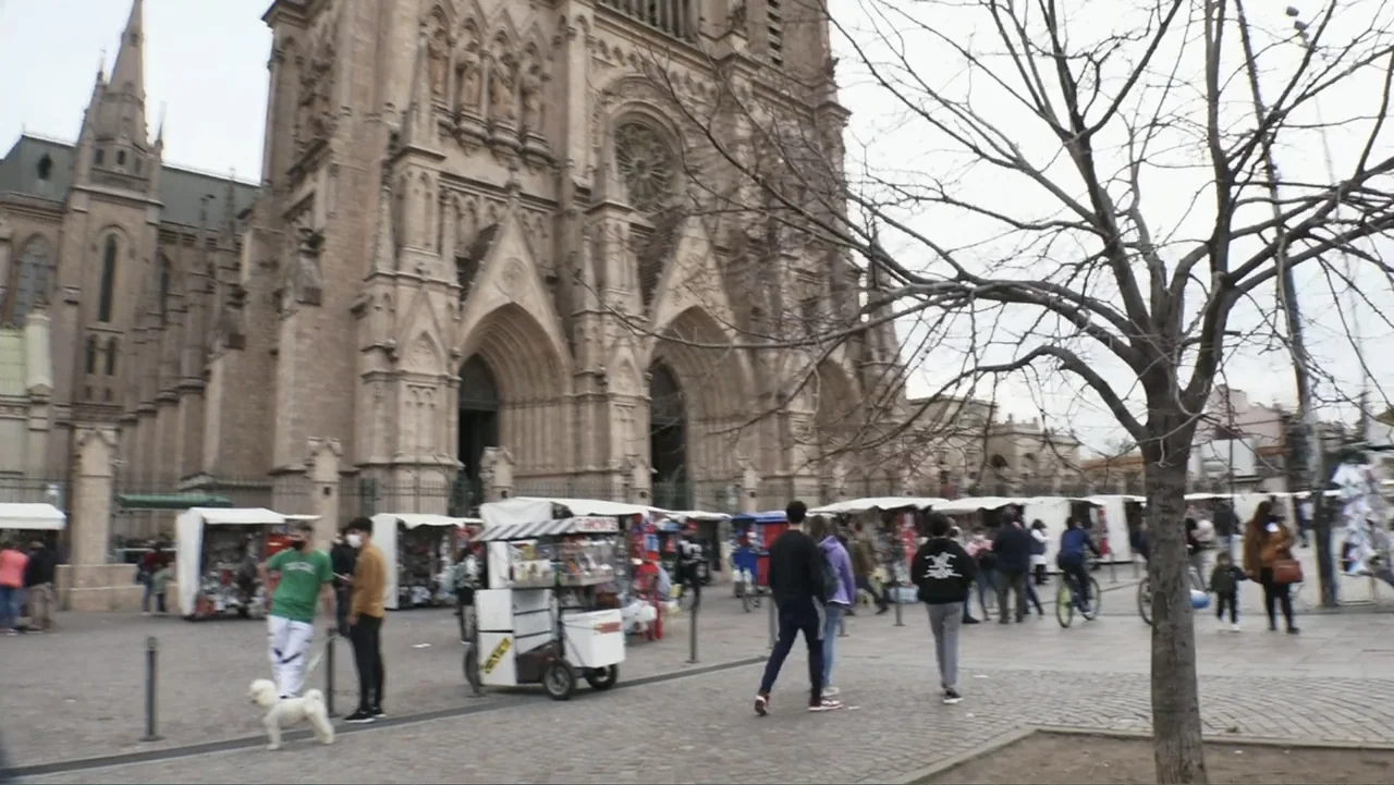 Saturdays Disorders (2022) movie still: wide — A stone gothic cathedral stands behind a wide plaza filled with white market stalls. Multiple people…