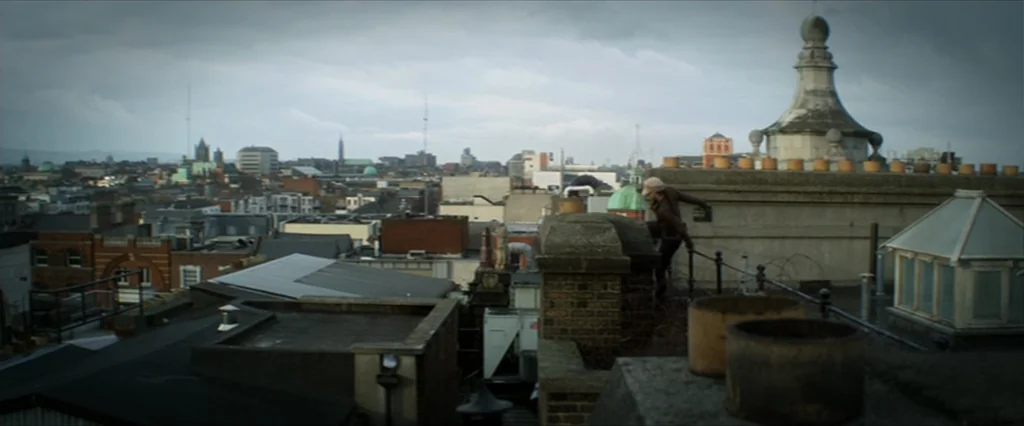 Haywire (2011) movie still: wide — A dense urban landscape extends to the horizon under a heavily clouded gray sky. A lone figure crouches on a…