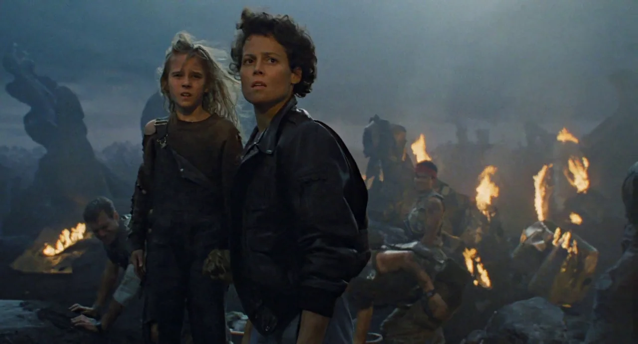 Aliens (1986) movie still: medium — A dark, smoke-filled rocky landscape at night features several small fires burning among wreckage. A woman in a…