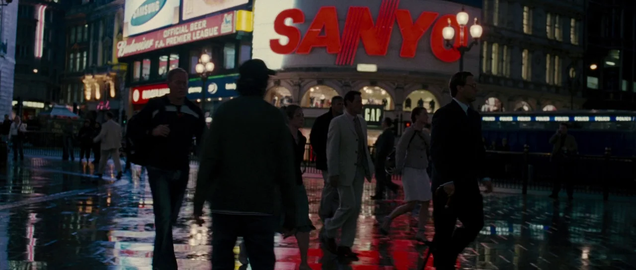 A Good Year (2006) movie still: wide — Pedestrians walk across a rain-slicked city square surrounded by large illuminated corporate advertisements.…
