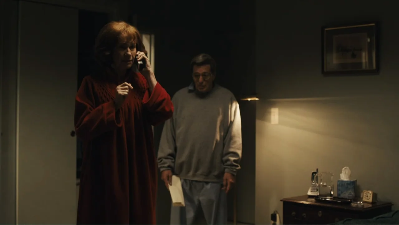 Paterno (2018) movie still: medium — A dim bedroom featuring a side table with a landline telephone and tissue box. A woman in a red robe speaks on…