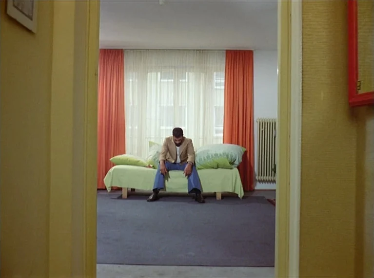 Ali: Fear Eats The Soul (1974) movie still: wide — A 1970s room with orange curtains and green bedspread is framed through a doorway. A man sits…