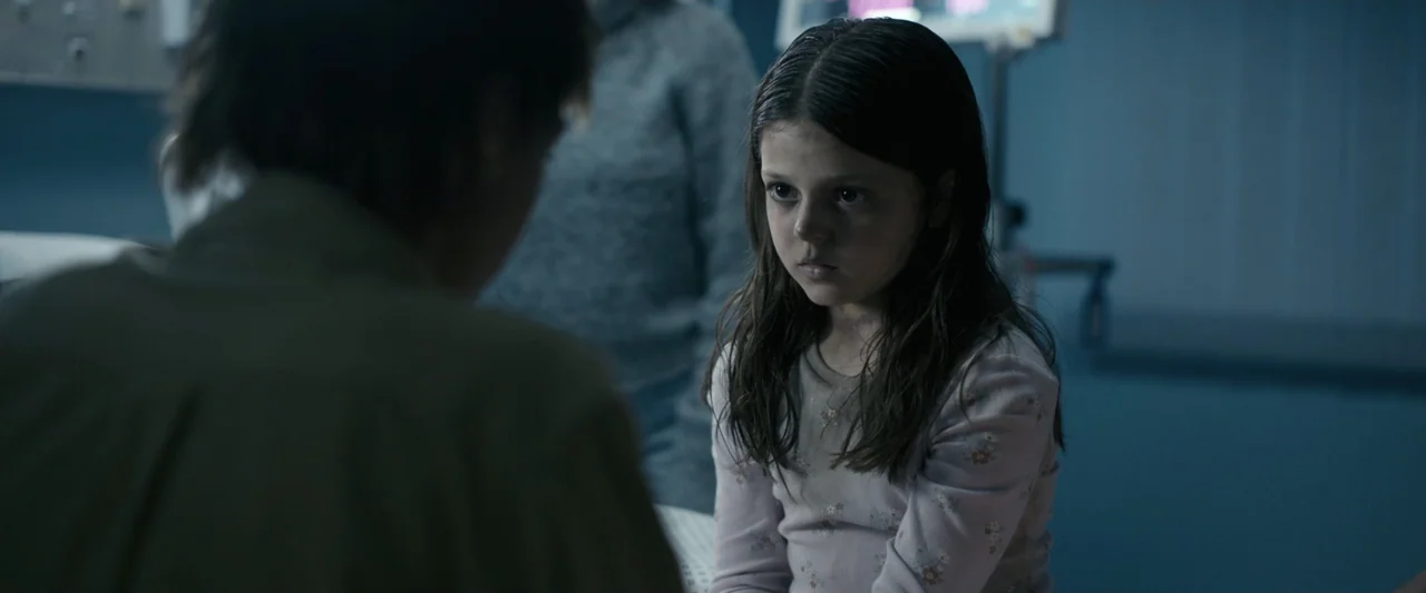 Talk to Me (2023) movie still: medium — A dimly lit hospital room with blue walls features medical equipment visible in the background. A young girl…