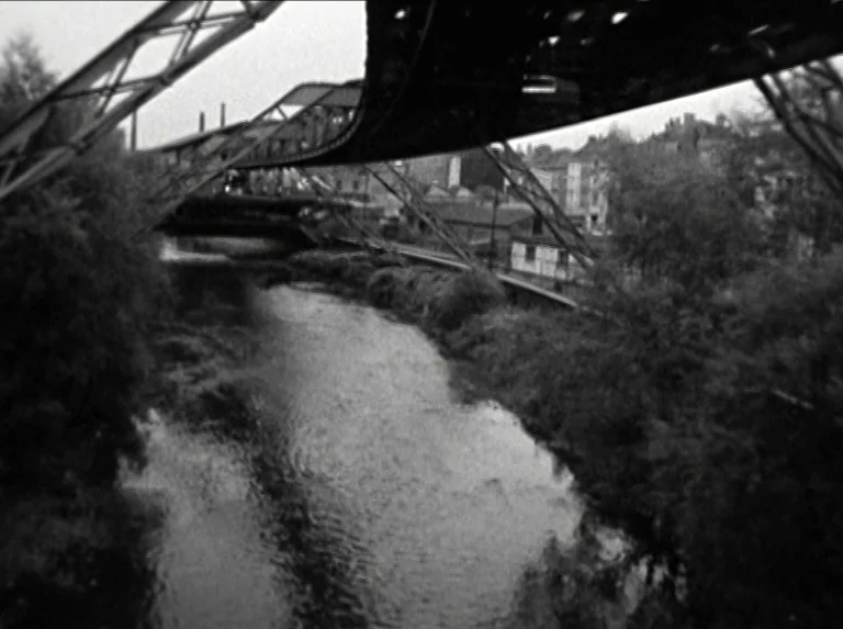 Alice in the Cities (1974) movie still: establishing — A dark metal suspended railway structure spans over a river flowing through a town. Sparse…