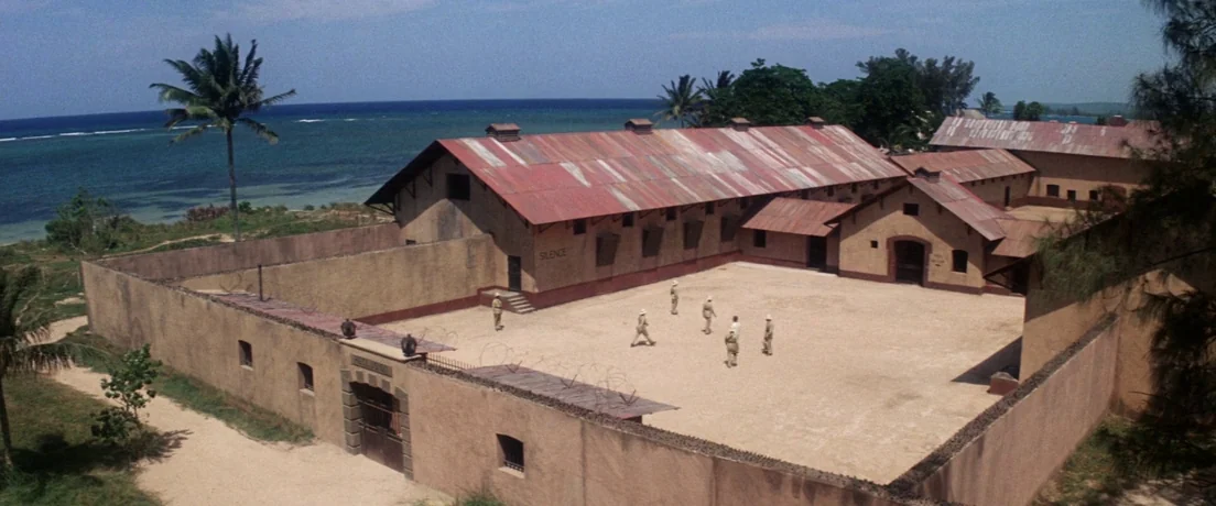 Papillon (1973) movie still: establishing — A fenced coastal compound features beige buildings with rusty corrugated metal roofs. Several soldiers…