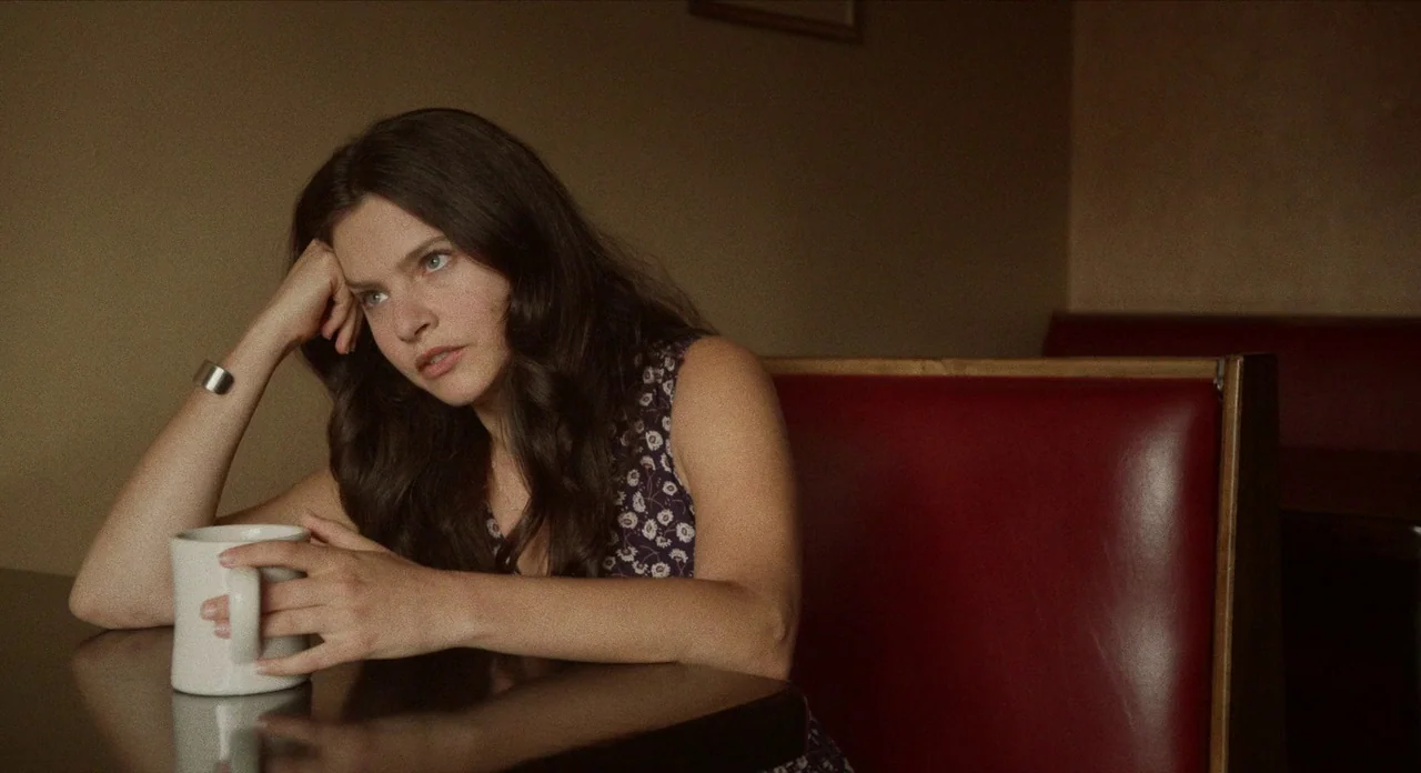 The Art of Self Defence (2019) movie still: medium — A diner booth with red vinyl seating and a dark table. A young woman sits at the table holding…