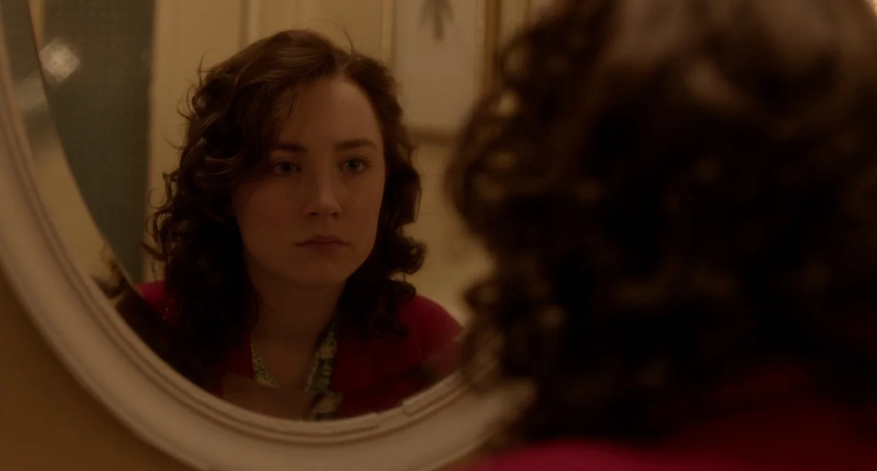 Brooklyn (2015) movie still: closeup — A woman looks at her reflection in an oval mirror mounted on a wall. Her face is centered in the reflection…