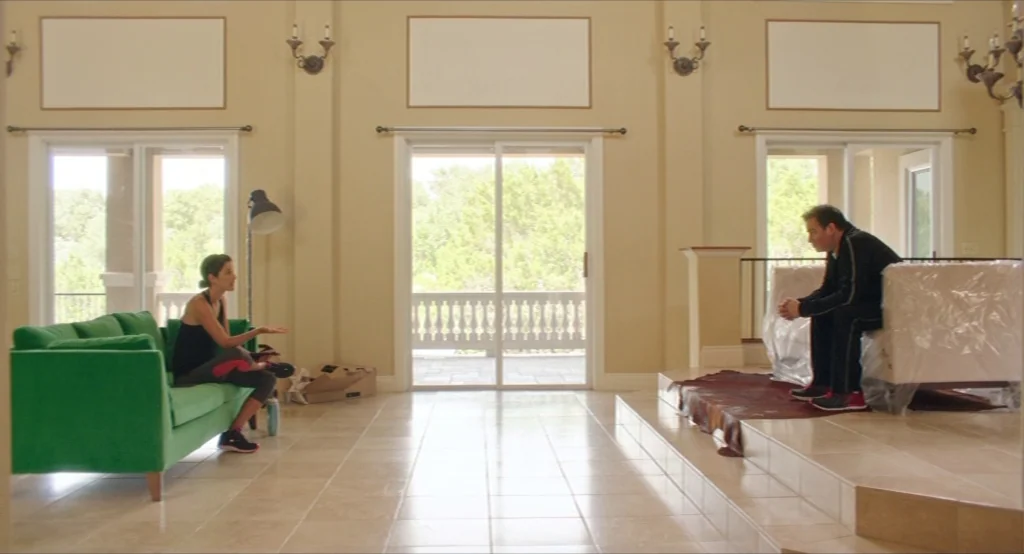 Results (2015) movie still: wide — A spacious room with tile floors and beige walls contains two large sofas. A woman on a green couch speaks to a…