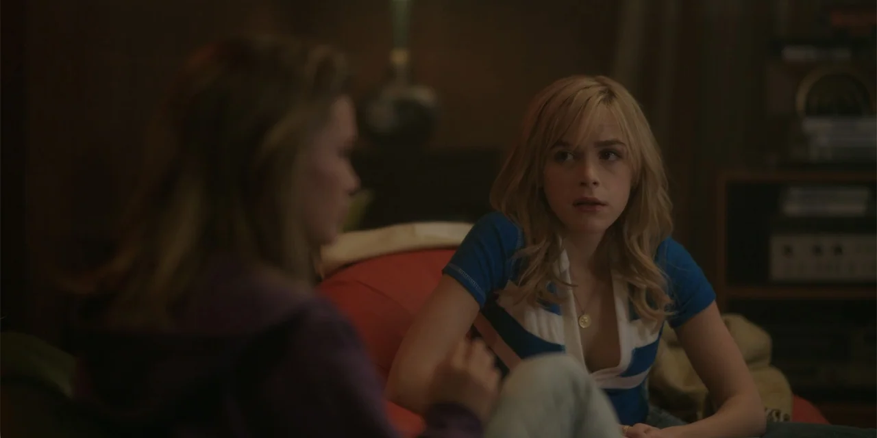 Totally Killer (2023) movie still: medium — A dimly lit room features a blonde woman sitting on a red furniture piece. She faces a second person in…