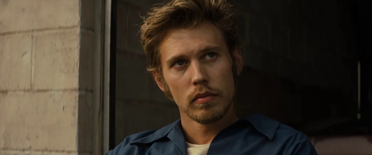 The Bikeriders (2023) movie still: closeup — A man stands inside a dimly lit garage space with cinder block walls. He gazes towards the right while…