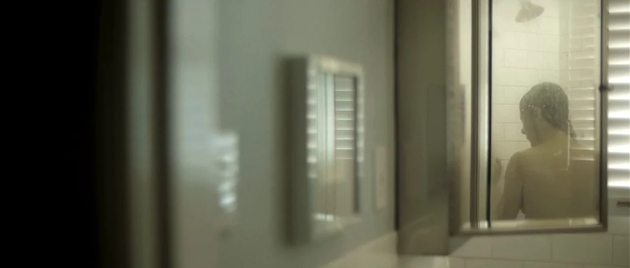 Upstream Color (2013) movie still: medium — A bathroom interior with white tiled walls and a partially open medicine cabinet mirror. A woman is…