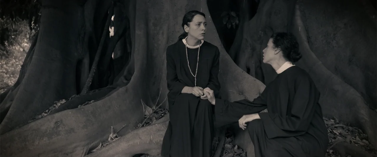 I Trust in You (2016) movie still: medium — Two women wearing religious habits sit among the large buttress roots of a tropical tree. One woman…