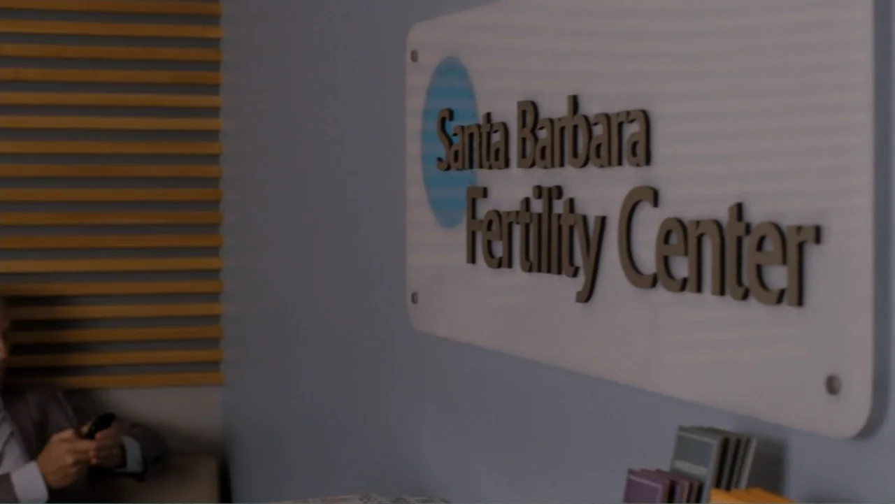 It's Complicated (2009) movie still: medium — A modern clinic waiting room features a prominent wall sign for a fertility center. A man wearing a…
