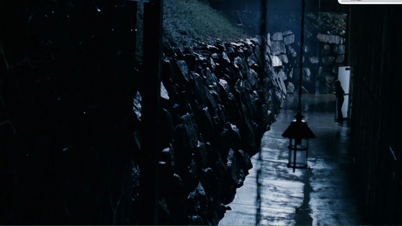 Chain Letter (2010) movie still: wide — A narrow exterior walkway is bounded by a wet stone retaining wall. A person stands in the distance at an…