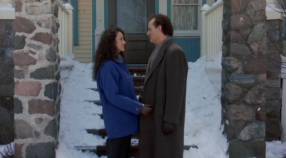 Groundhog Day (1993) movie still: medium — A residential house front with stone pillars and steps covered in deep snow. A man and woman stand facing…