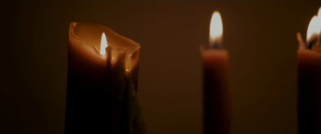 MacGruber (2010) movie still: closeup — Multiple tall candles burn in a darkened room, casting limited light on the wax. The flames glow brightly…