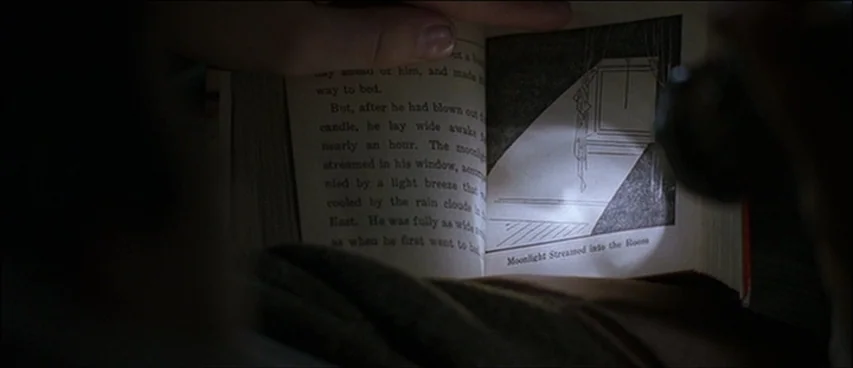 Road To Perdition (2002) movie still: closeup — An open book rests on a person's lap in a dark room. A focused beam of light reveals text and a…