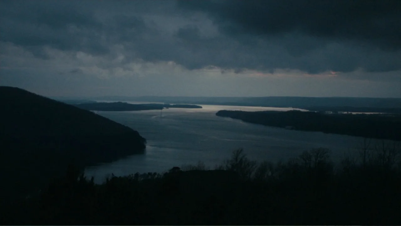 Alabama Snake (2020) movie still: establishing — A wide river winds through a dark valley beneath a heavy cloud-covered sky. Sparse silhouettes of…