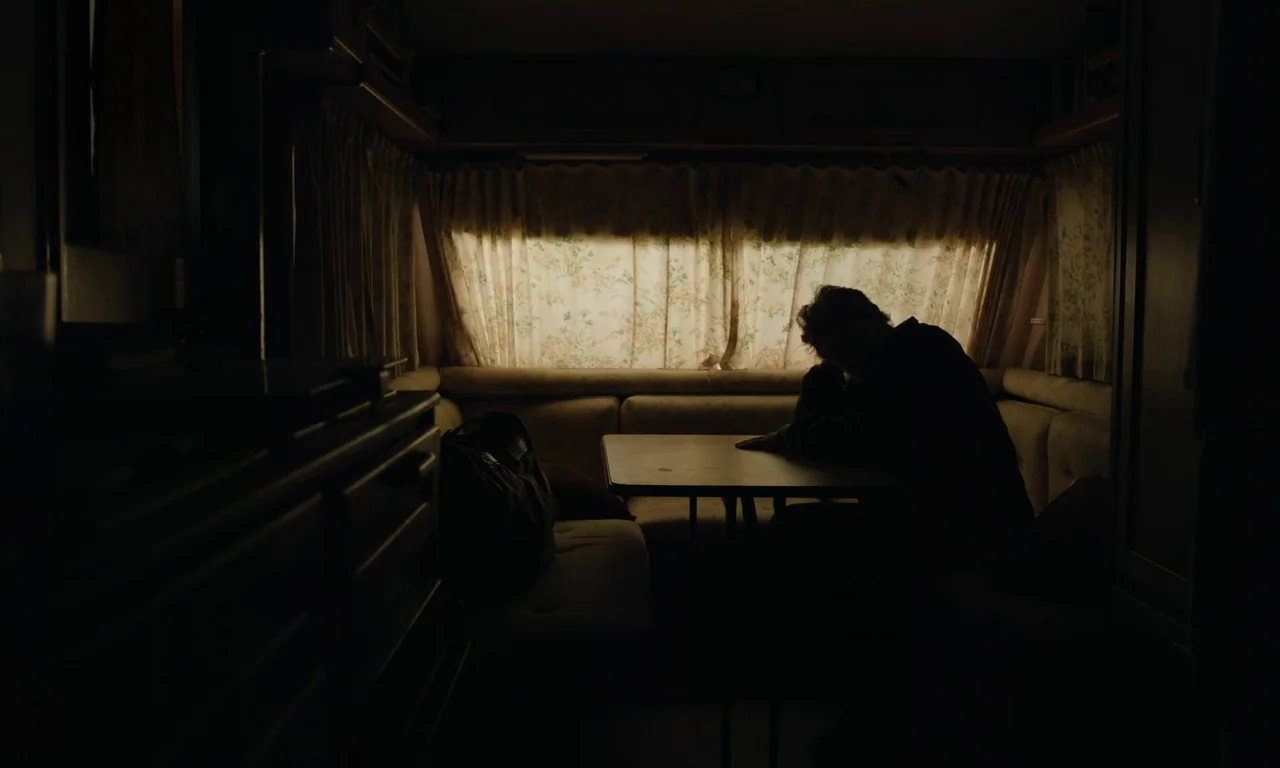 Close Your Eyes (2023) movie still: medium — A dark camper interior features a table and benches beneath a curtained window. A man sits silhouetted…