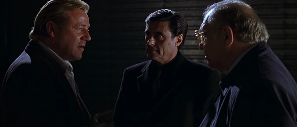 Sexy Beast (2000) movie still: medium — A dark indoor space with horizontally ribbed metal walls serving as a backdrop. Three men stand in a cluster…