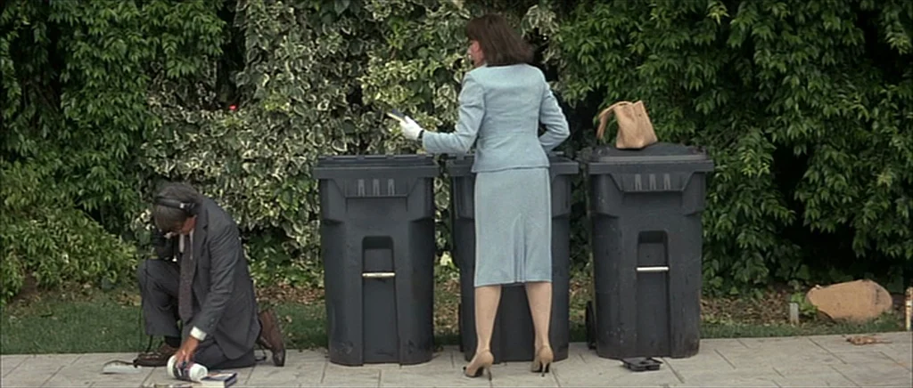 I Heart Huckabees (2004) movie still: wide — A sidewalk paved with light grey stone sits in front of a dense hedge. A kneeling man wearing a suit…