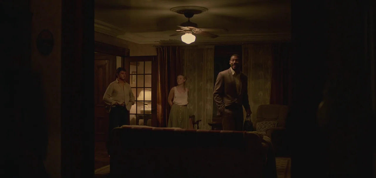 Foe (2023) movie still: wide — A dimly lit room with heavy curtains and a central ceiling fan light. Three people stand in the room while looking…