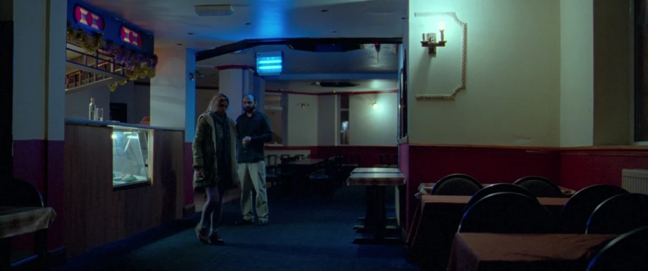 Catch Me Daddy (2014) movie still: wide — A dimly lit, carpeted dining room contains scattered tables and chairs under blue light. A man and woman…