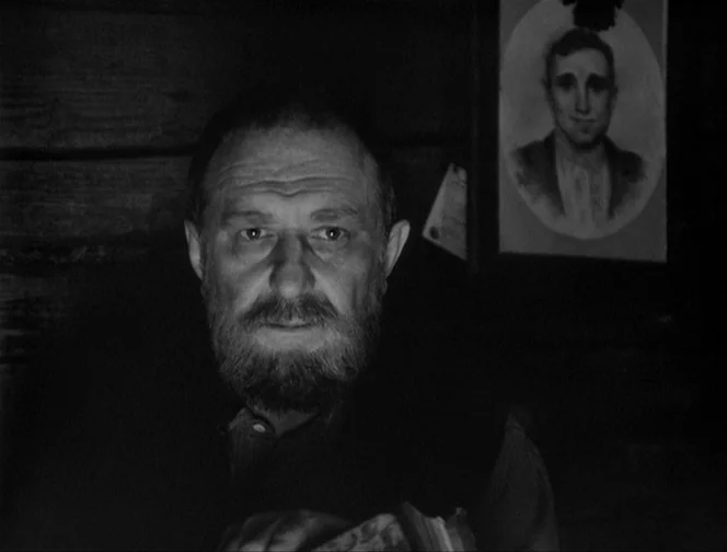 The Ascent (1977) movie still: closeup — A dimly lit room with dark wood walls and a framed portrait on the wall. A middle aged man with a full…
