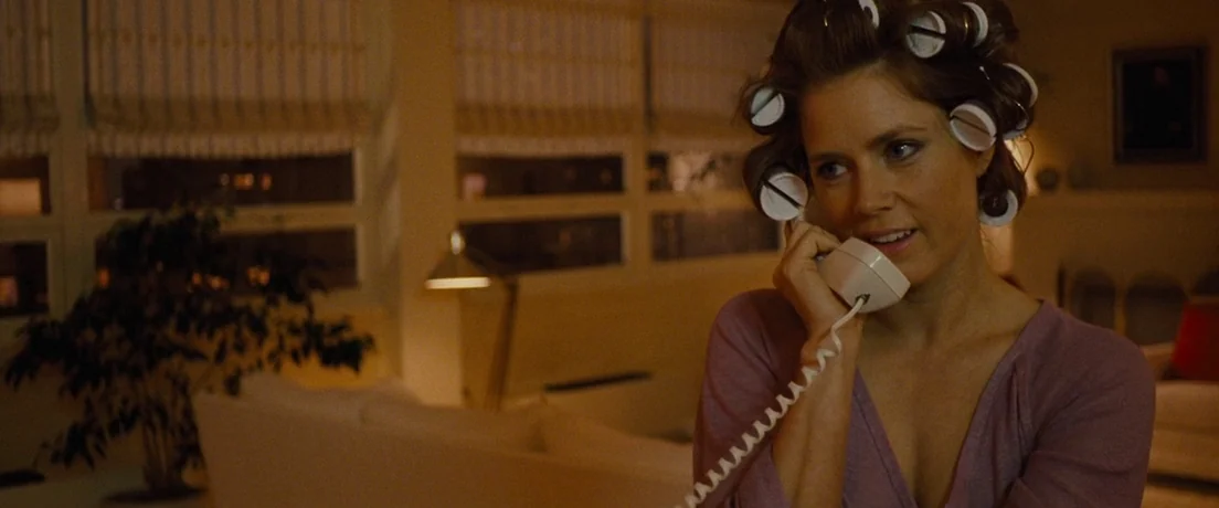 American Hustle (2013) movie still: medium — A dimly lit room with large windows showing a dark nighttime cityscape outside. A woman with hair…