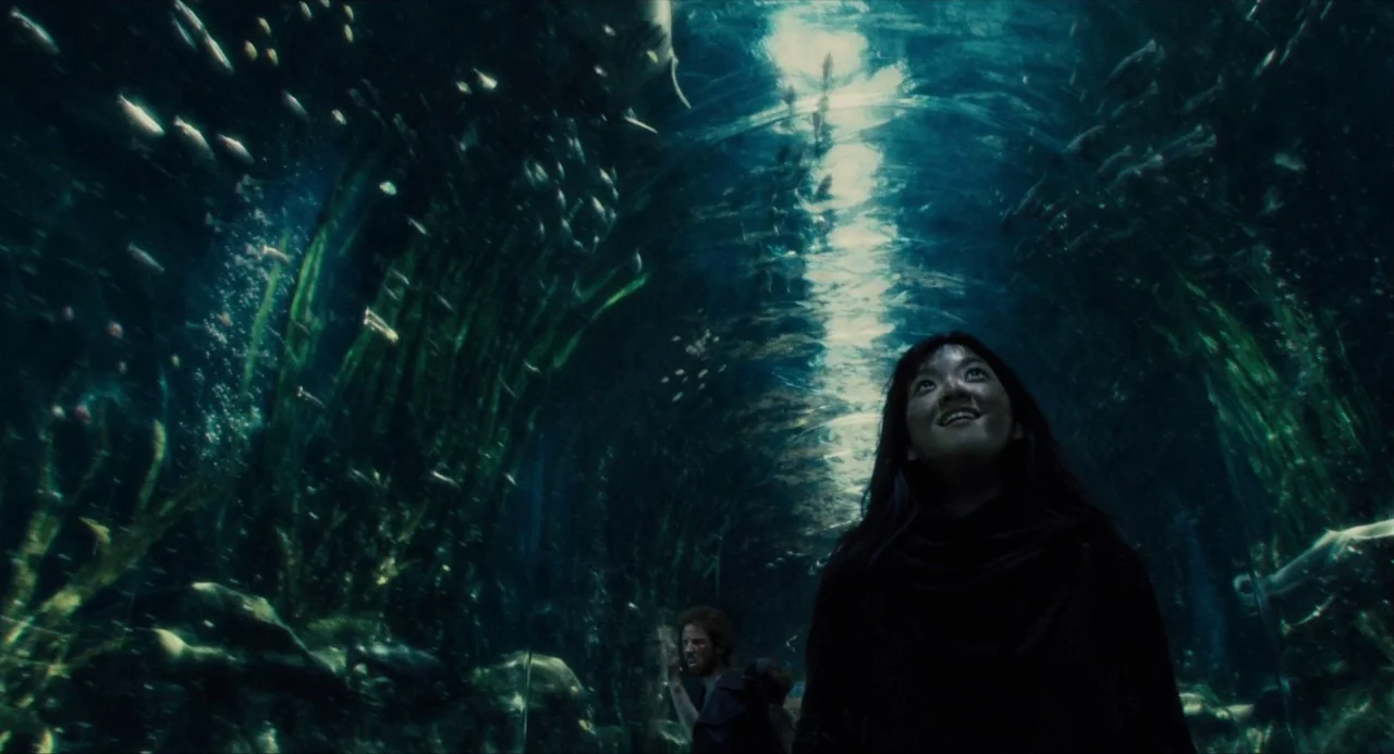 Snowpiercer (2013) movie still: medium — A glass tunnel passes through a dark, deep-water aquarium exhibit filled with swimming fish. A woman walks…