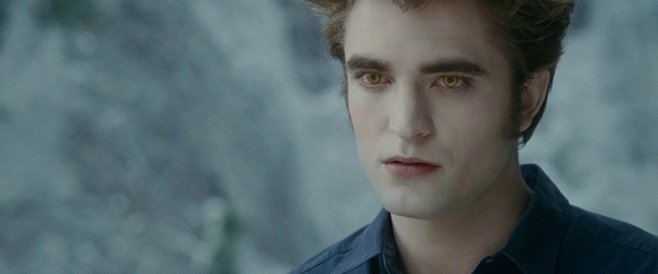 The Twilight Saga: Eclipse (2010) movie still: closeup — A man with pale skin and amber eyes stands against a blurred gray rocky backdrop. He…